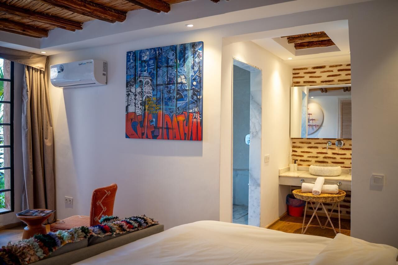 A hotel room with a bed, a wall-mounted air conditioner, a colorful abstract painting, a bathroom with a brick wall, and a small round table with towels.