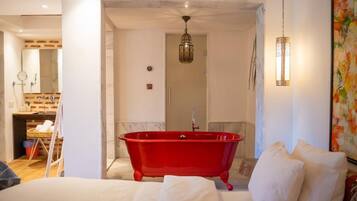 Une pièce avec une baignoire sur pieds rouge, un lit avec du linge de maison blanc, un luminaire suspendu et un tableau au mur.
