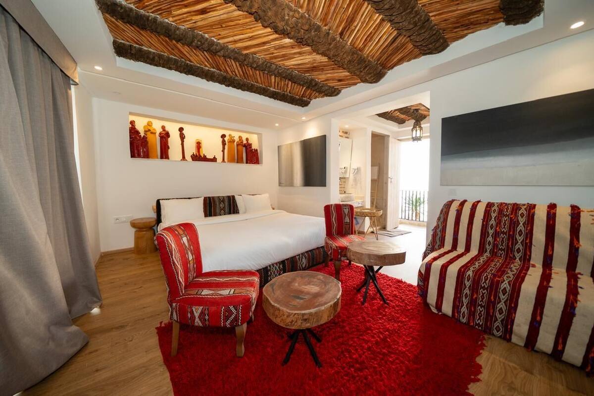 A hotel room with a bed, two chairs, a small table, a red rug, and a unique ceiling design.