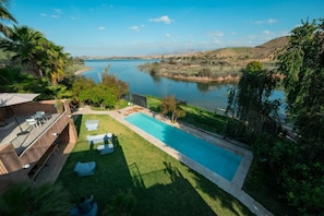 Piscina con vistas a un lago, rodeada de vegetación y una terraza con mobiliario exterior.