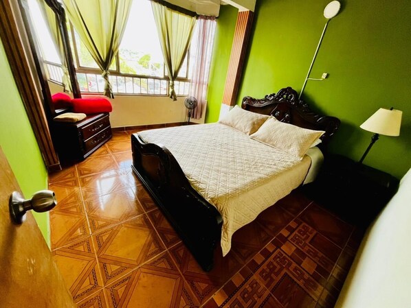 A bedroom with a bed, a dresser, a nightstand, a lamp, and a window with curtains.