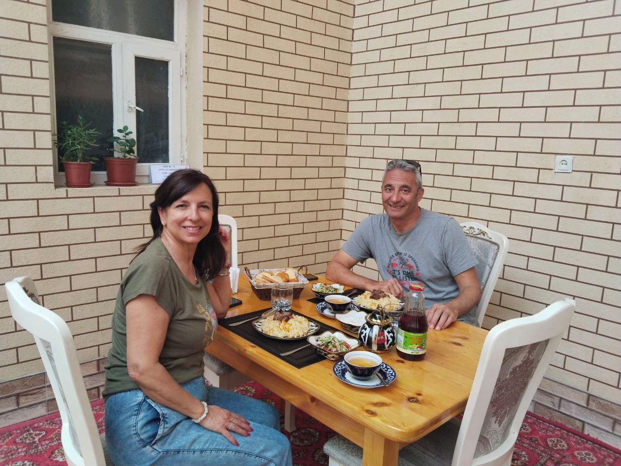 Two people seated at a wooden table with various dishes and drinks, in a room with brick walls and white chairs.
