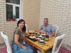 Deux personnes sont assises à une table en bois, devant divers plats et boissons, dans une pièce aux murs en briques et aux chaises blanches.