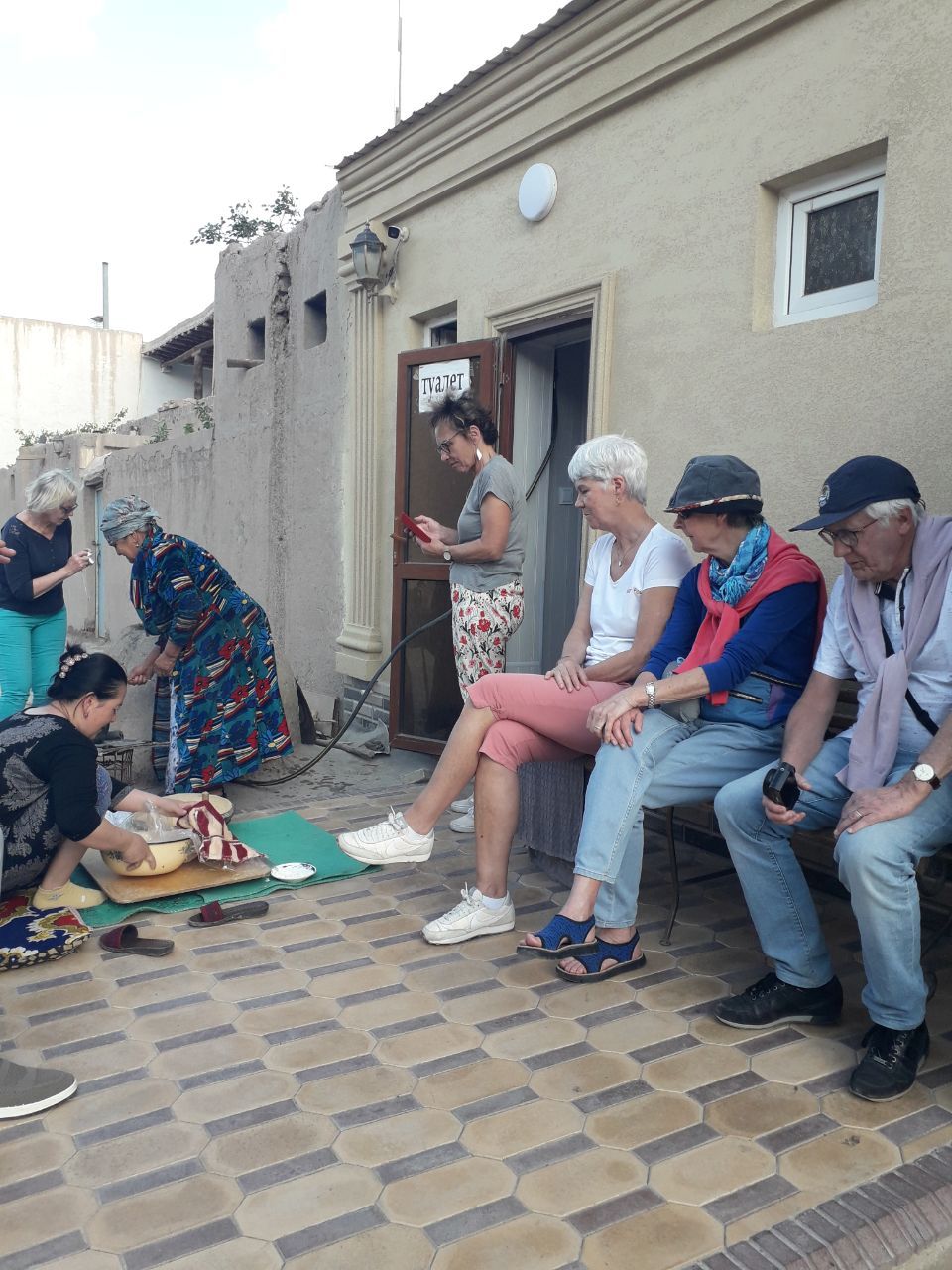 A group of people sitting and standing on a tiled patio area with a building in the background.