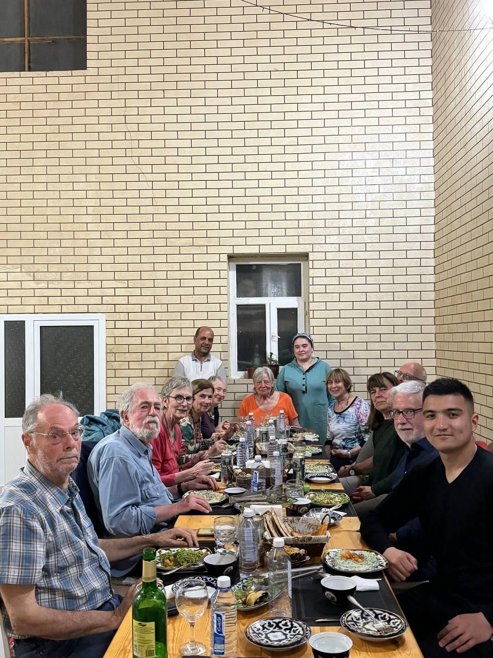 Un groupe de personnes est assis autour d’une longue table, devant divers plats et boissons, dans une pièce aux murs de briques et dotée d’une fenêtre.