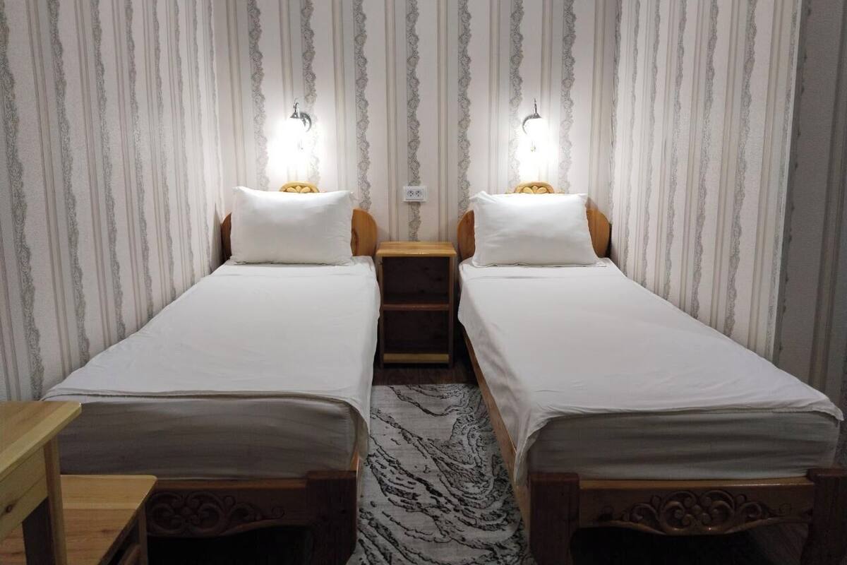 A room with two single beds, wooden headboards, a small bedside table, and a patterned rug.