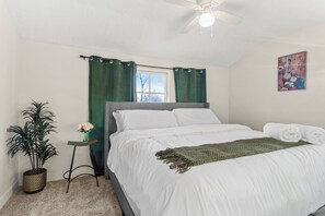 Une chambre à coucher avec un lit, un ventilateur de plafond, une fenêtre avec des rideaux, une plante en pot et une petite table sur laquelle se trouve un vase.