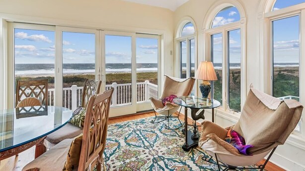 Et værelse med et glasbord, stole, en lampe og store vinduer med udsigt til stranden.