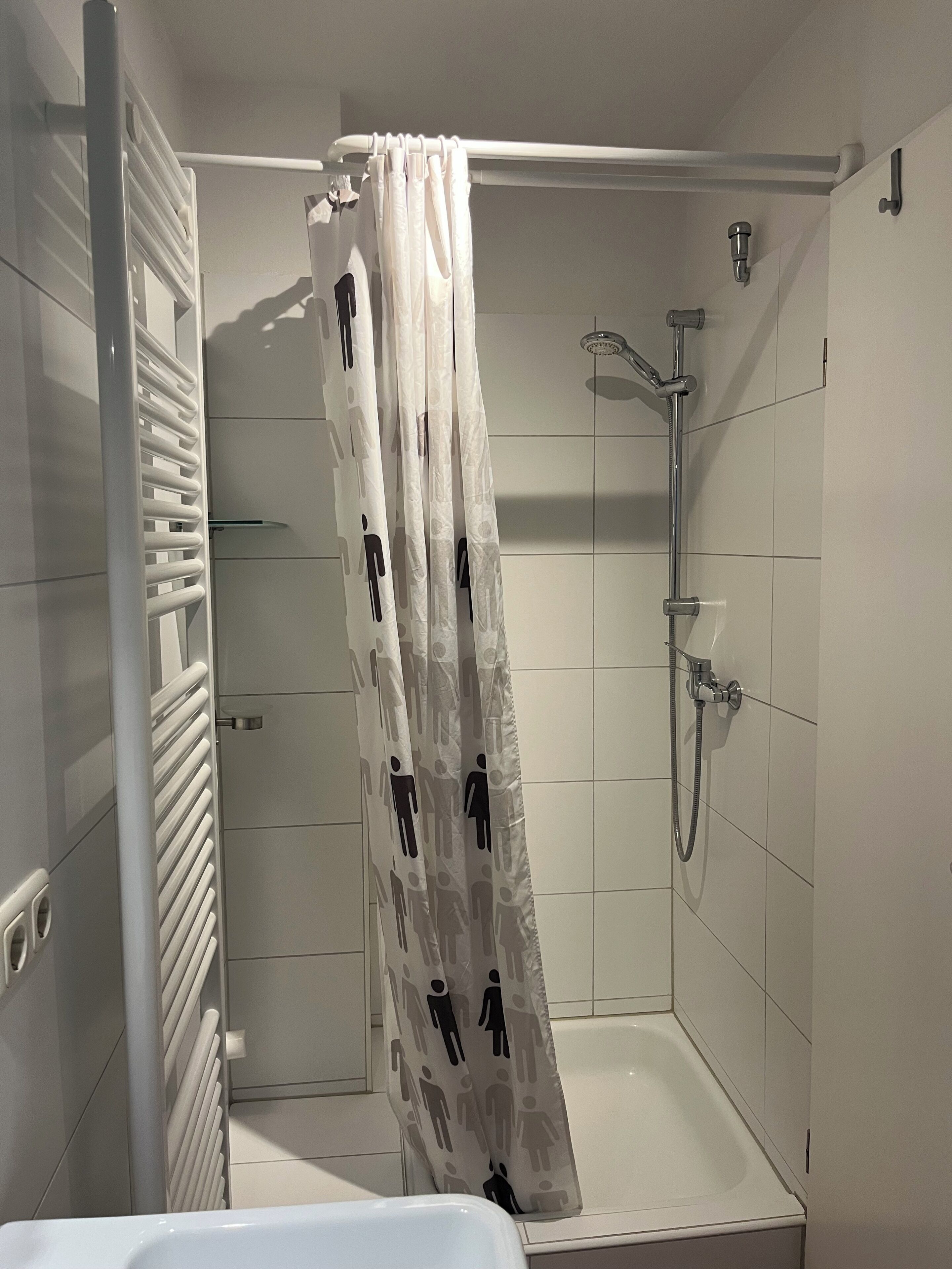 Eine Dusche mit weiß gefliesten Wänden, einer Duschbrause und einem Vorhang mit einem Muster aus schwarzen Figuren.