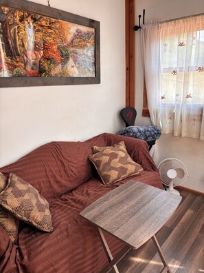 A living room with a brown sofa, patterned pillows, a small table, a fan, and a painting on the wall.