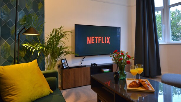 Un salón con una televisión mostrando 'NETFLIX', un sofá, una mesita de centro con un croissant y jugo, y una planta.