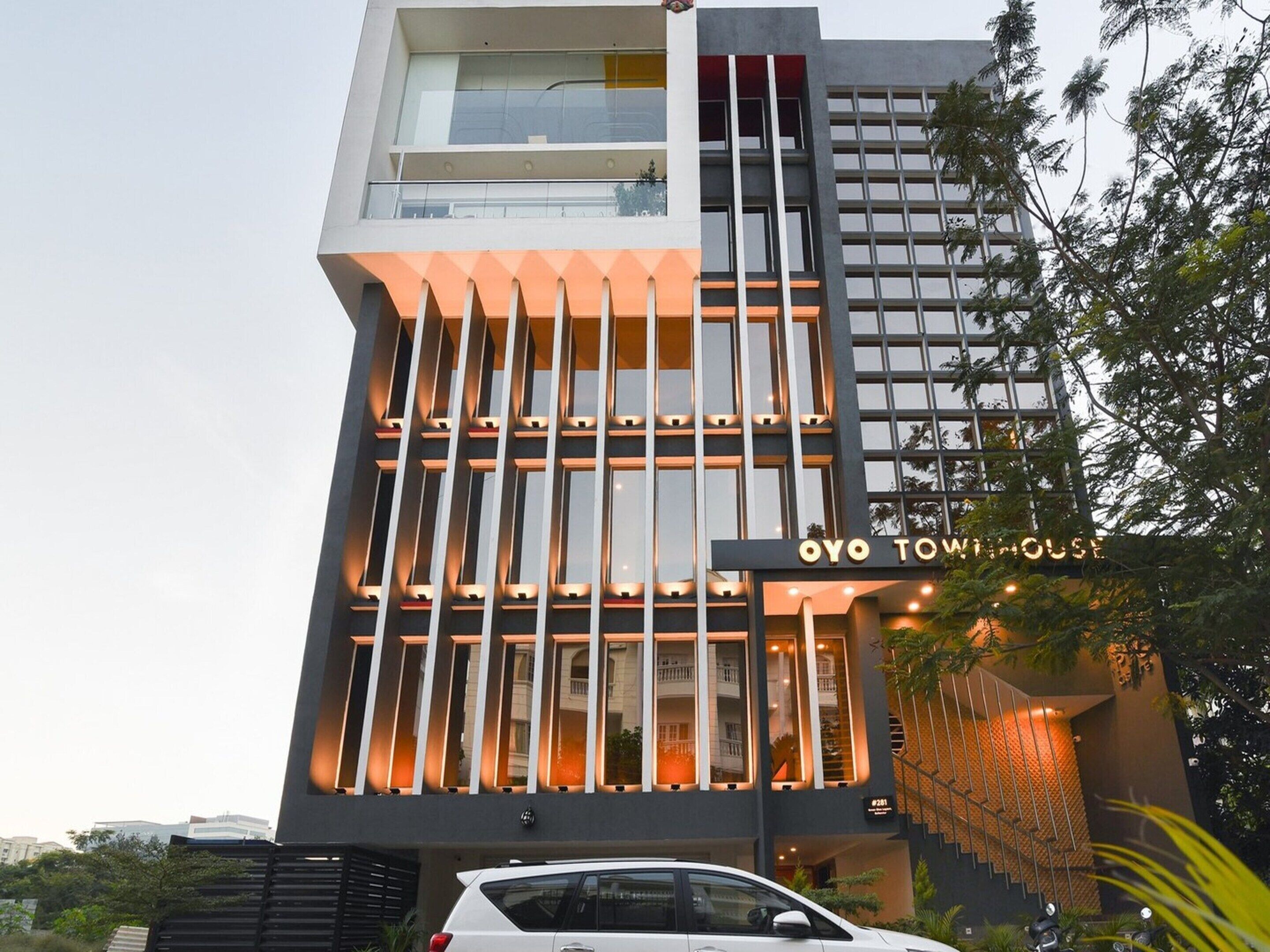Egy modern hotel, feltűnő, függőleges vonalvezetésű építészettel, egy „OYO TOWING HOUSE” felirattal ellátott táblával, és egy lépcsővel, amely a bejárathoz vezet.