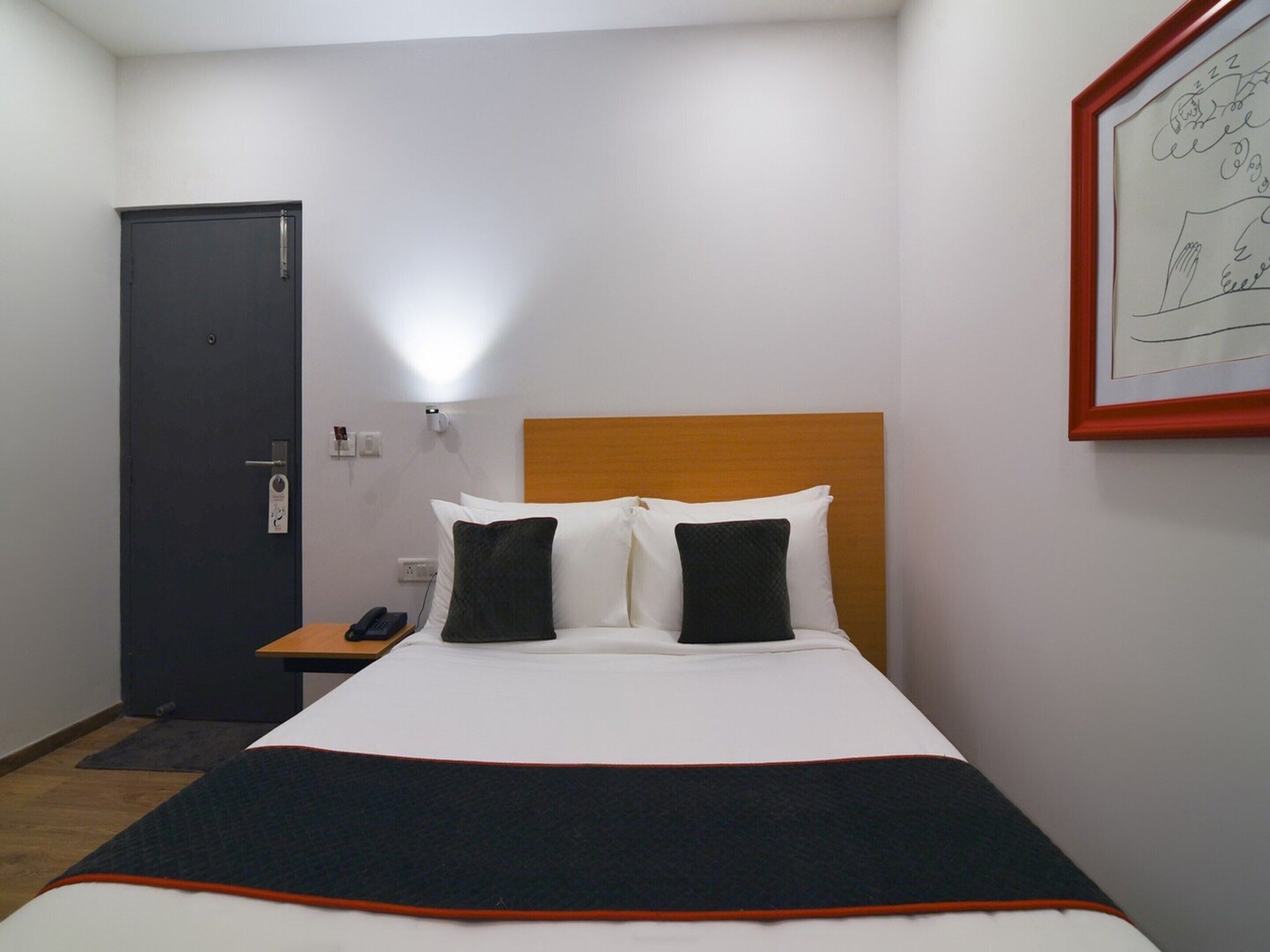 A hotel room with a bed, a bedside table with a phone, a door, and a wall-mounted light fixture.