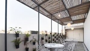 Une terrasse sur le toit, avec une table ronde et des chaises en métal blanc, entourée de plantes en pot.