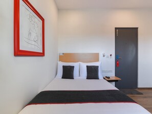 Ein Hotelzimmer mit Bett, Nachttisch, Tür und einem gerahmten Bild an der Wand.