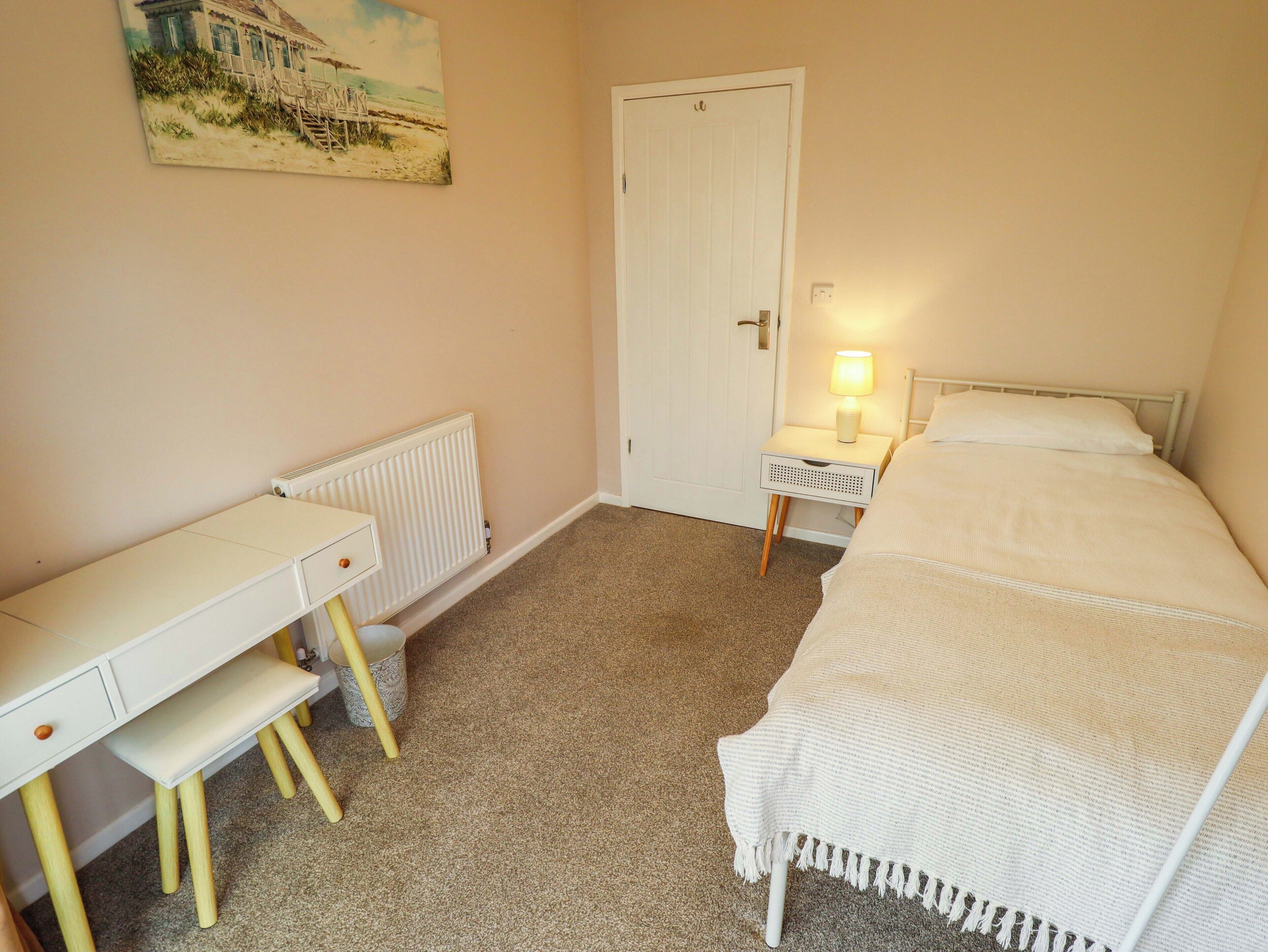 A bedroom with a bed, a desk, a chair, a radiator, and a nightstand with a lamp.