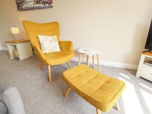 Ein Zimmer mit einem gelben Sessel, passendem Hocker, einem kleinen weißen Tisch, einer Lampe und einem Fernseher auf einem Schrank.