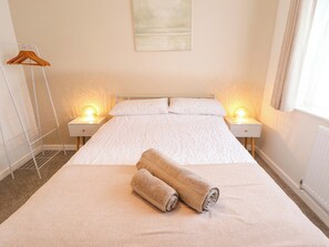 A neatly made bed with two rolled towels, two bedside lamps, a white bedside table, a wooden hanger, and a radiator. - Field View (North Walsham)