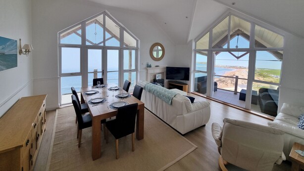Ein großzügiger Wohnbereich mit Esstisch, Sofas und großen Fenstern mit Blick auf einen Strand.