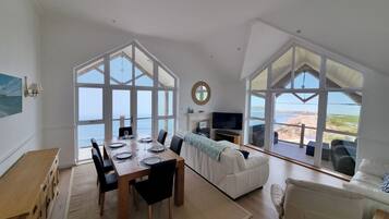 Ampla sala de estar com mesa de jantar, sofás e grandes janelas com vista para a praia.
