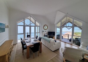 A spacious living area with a dining table, sofas, and large windows overlooking a beach. - 11 Thurlestone Rock (Kingsbridge)
