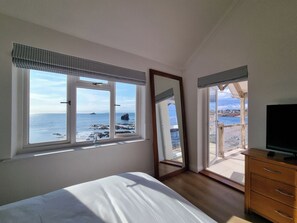 Ein Schlafzimmer mit einem Bett, einem Schminktisch, einem Fernseher und einem großen Fenster mit Blick auf das Meer und ein Schiff.