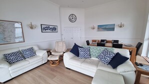 Ein Wohnzimmer mit zwei weißen Sofas, einem beigefarbenen Sessel, einem Couchtisch aus Holz und einem Essbereich mit Holztisch und Stühlen.