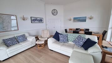 A living room with two white sofas, a beige armchair, a wooden coffee table, and a dining area with a wooden table and chairs.