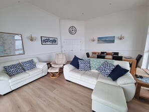 Ein Wohnzimmer mit zwei weißen Sofas, einem beigefarbenen Sessel, einem Couchtisch aus Holz und einem Essbereich mit Holztisch und Stühlen.