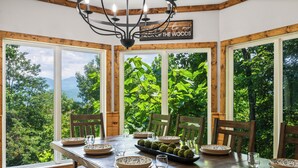 A dining area with a wooden table set for four, large windows with a view of trees and mountains, and a chandelier.