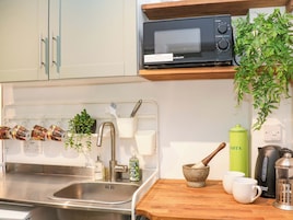 Una cocina con un lavamanos de acero inoxidable, un microondas, un recipiente verde para pasta, una cafetera y una planta colgante.