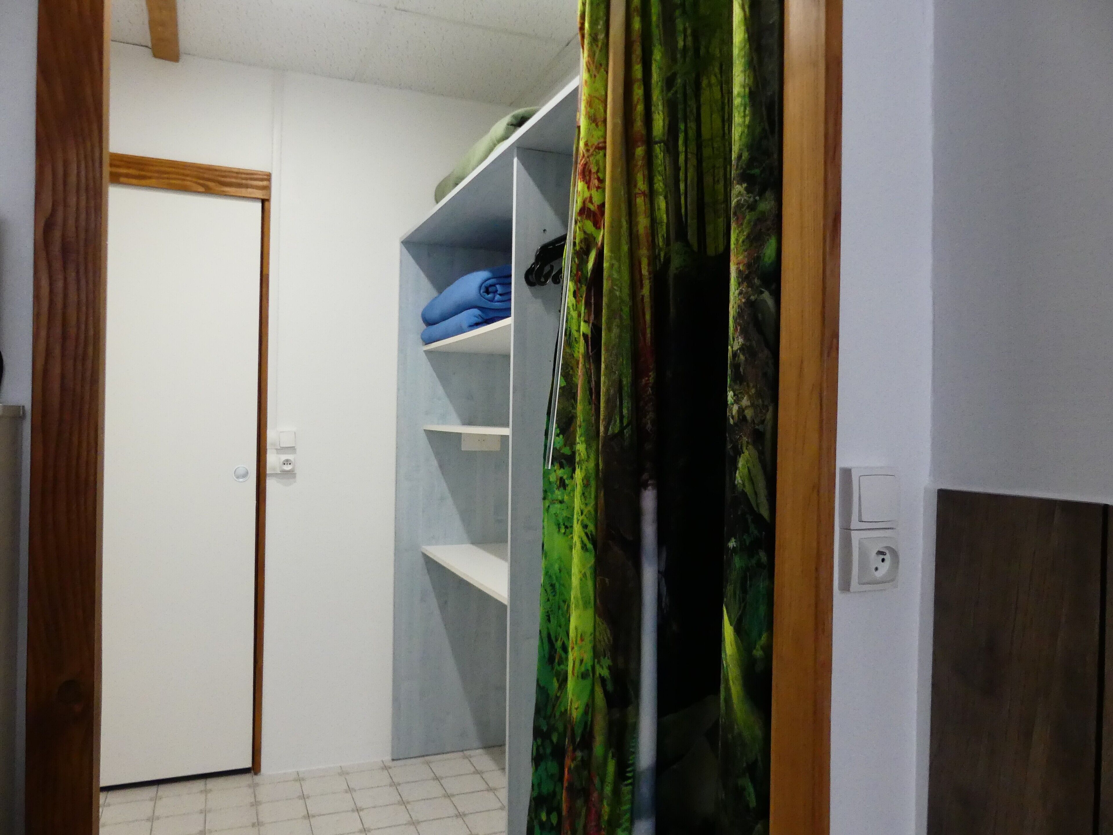 Een kamer met een witte deur, een inbouwkast met planken, en een gordijn met een bosmotief.