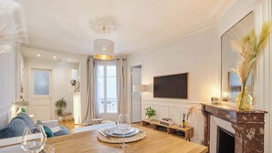 A dining area with a wooden table, chairs, and a wine glass. There is a fireplace, a television mounted on the wall, and a window with curtains. - Charming apartment in the center of Paris (Paris)