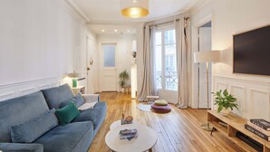 A living room with a blue sofa, a round coffee table, a TV, and a large window with curtains. - Charming apartment in the center of Paris (Paris)