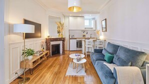 A living room with a sofa, a coffee table, a floor lamp, a fireplace, and a kitchen area in the background. - Charming apartment in the center of Paris (Paris)