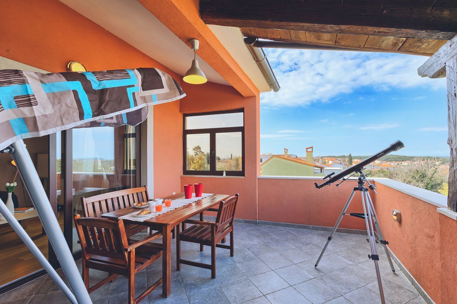 En balkon med et teleskop, et bord med stole og udsigt over bybilledet.