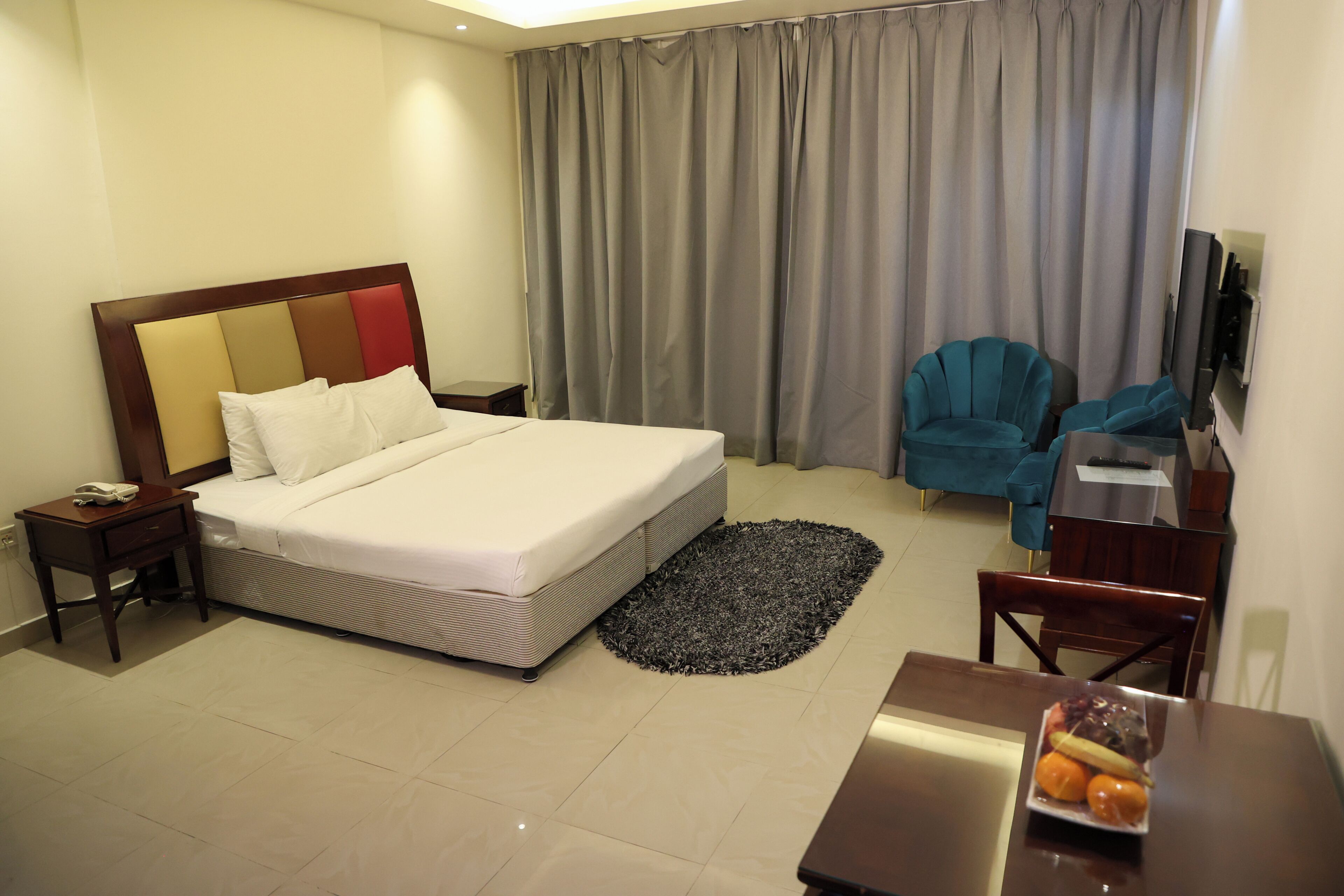 A hotel room with a bed, a desk with a chair, a television, and a dining table with fruit.