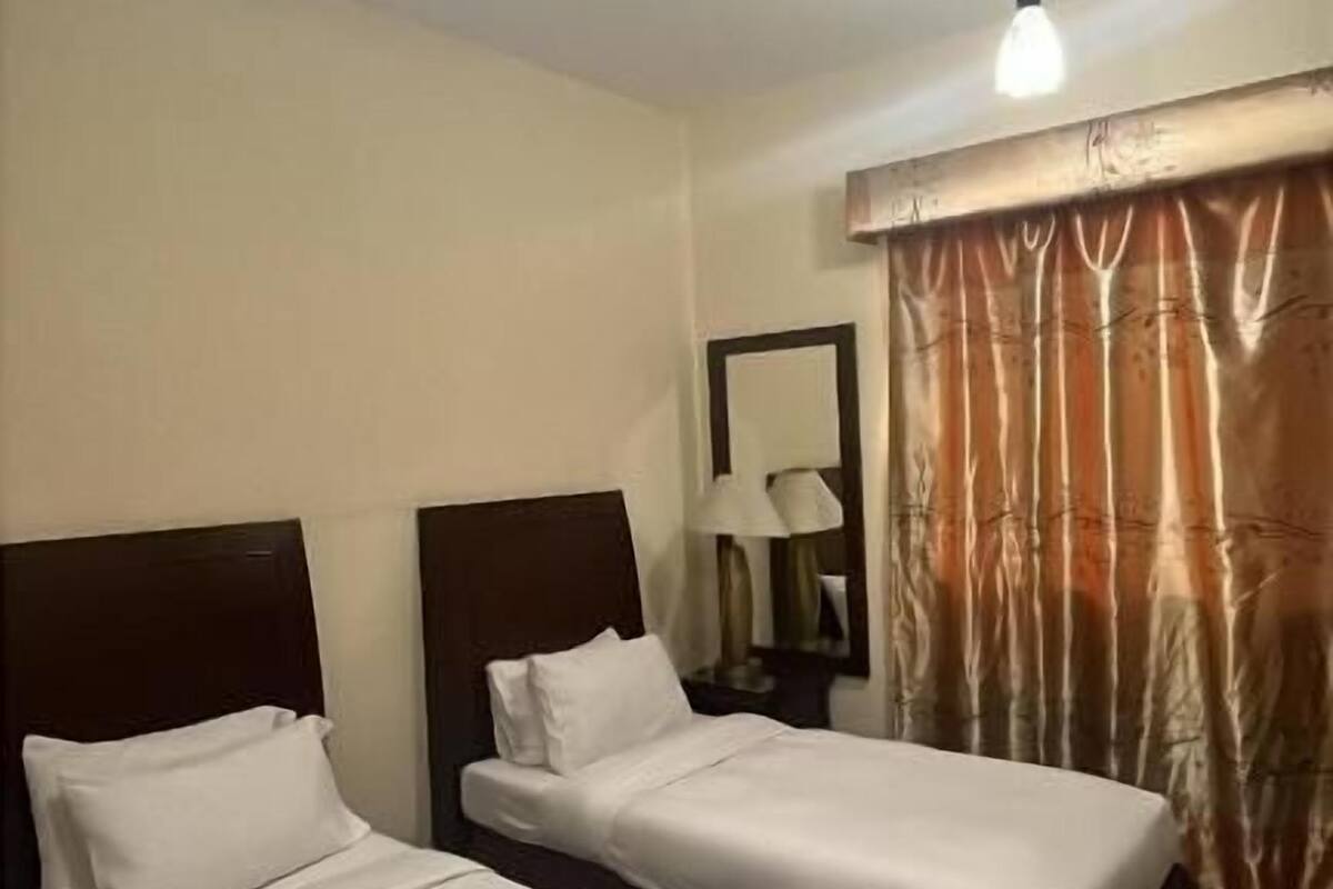 A hotel room with two single beds, a nightstand with a lamp, a mirror, and a window with curtains.
