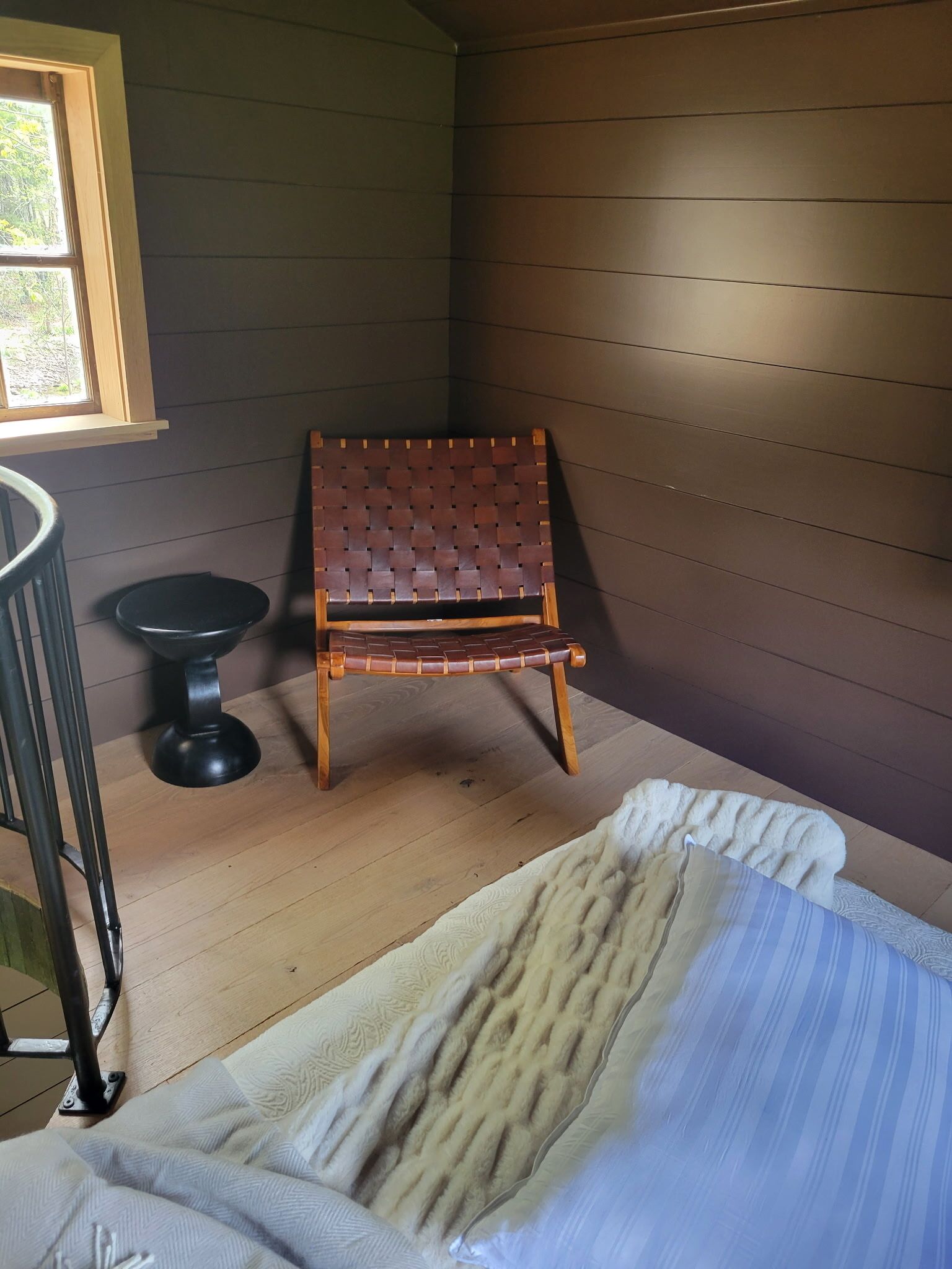 A room with a wooden floor, a black metal chair, a small round table, and a bed with a striped blanket.