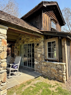 Une cabane rustique aux murs de pierre, au toit en bois et à la terrasse couverte, dotée de chaises.