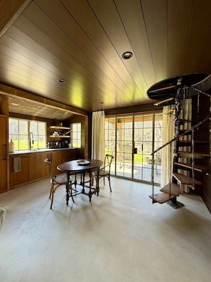 Une cuisine rustique avec des meubles en bois, une table à manger entourée de chaises, un escalier en colimaçon et de grandes fenêtres.