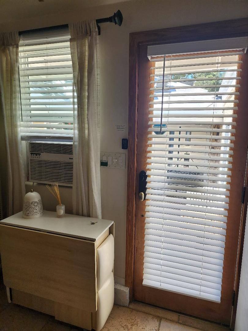 A room with a wooden door, a window with blinds, a small table with a vase and a decorative item, and a wall-mounted air conditioning unit.