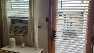 A room with a wooden door, a window with blinds, a small table with a vase and a decorative item, and a wall-mounted air conditioning unit.