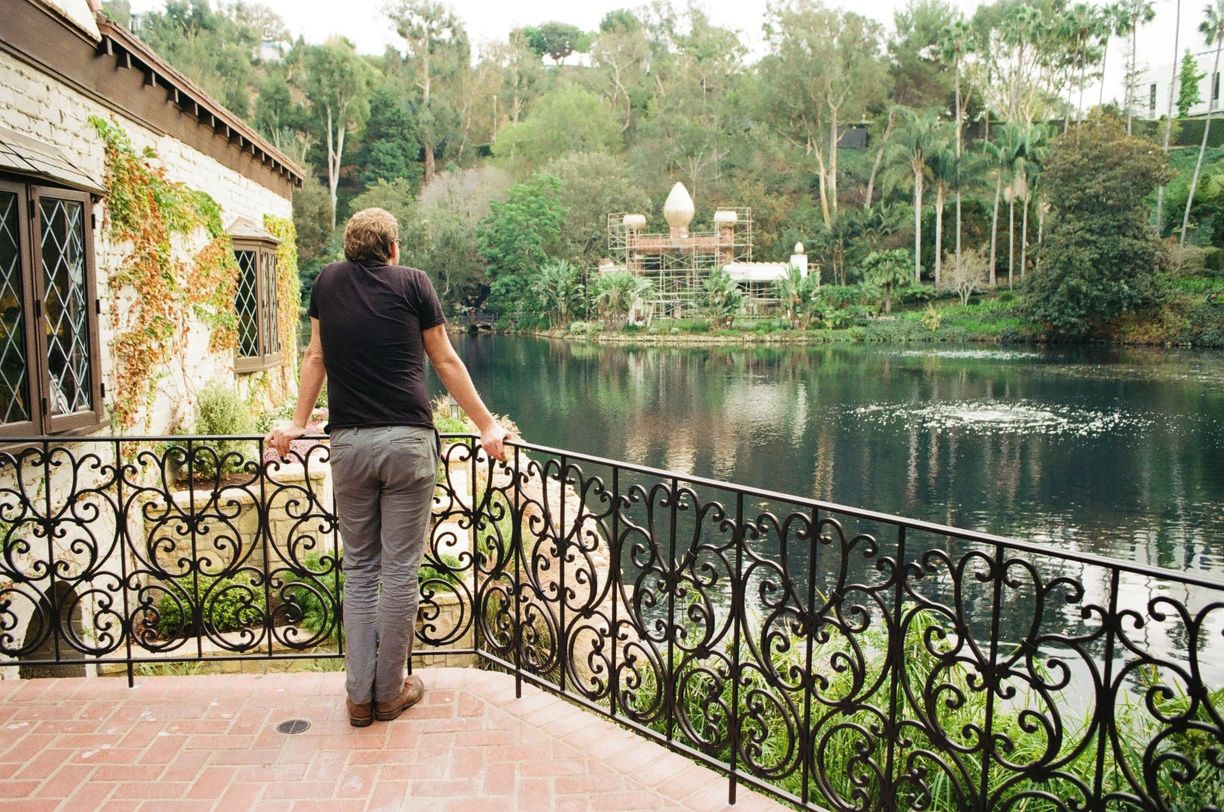 A person standing on a balcony with a view of a lake, a building with ivy, and a structure with a dome-like roof in the background.
