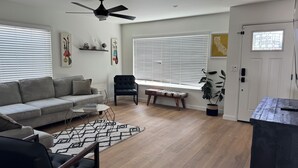 A living room with a sofa, a chair, a coffee table, a floor lamp, a ceiling fan, and a window with blinds.