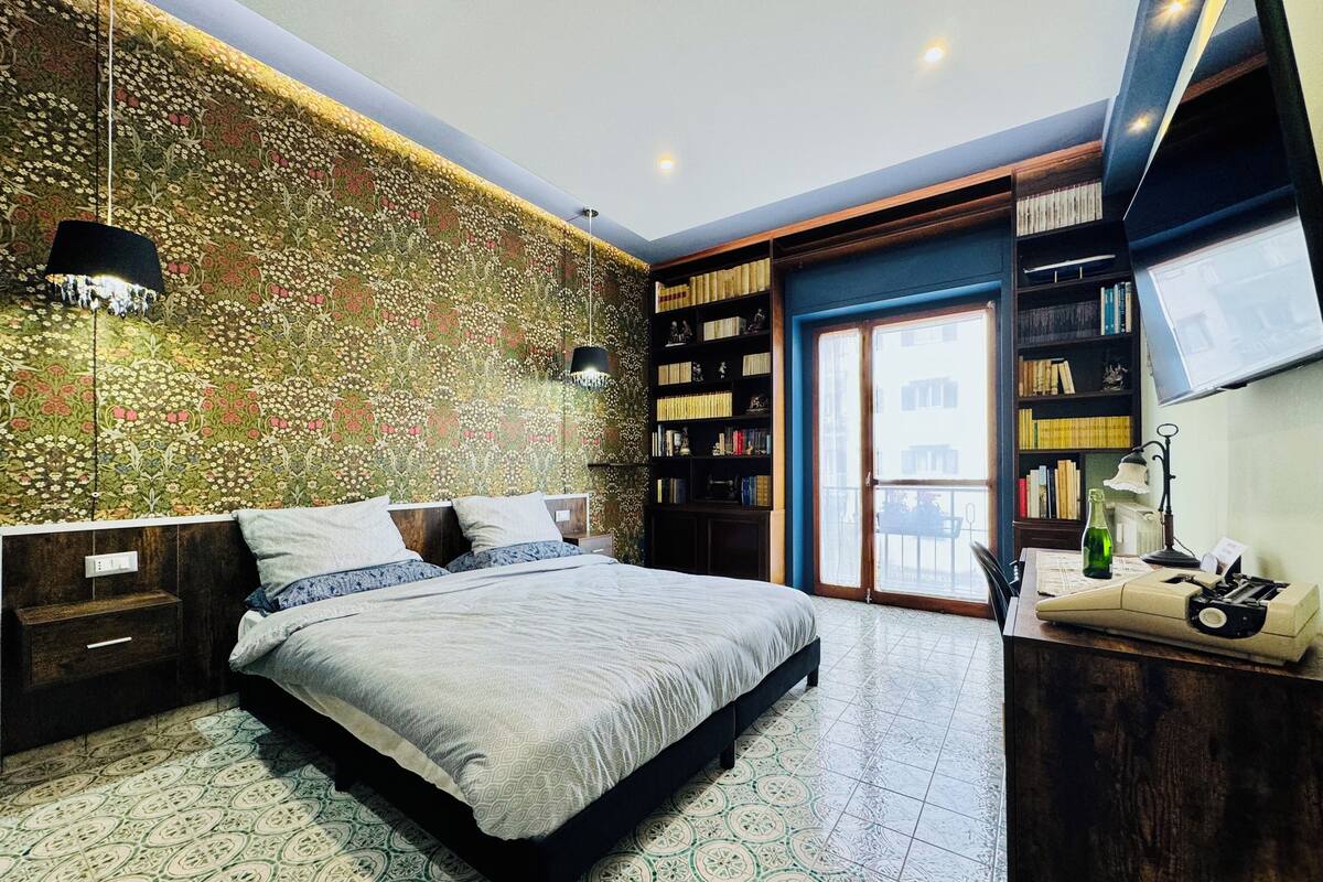 Un dormitorio con papel tapiz estampado, una cama con colcha estampada, una estantería y un escritorio con lámpara.