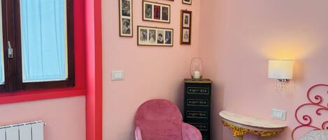 A room with a pink wall, a window with curtains, a pink chair, a black dresser with drawers, a small table with a lamp, and a bed with patterned bedding.