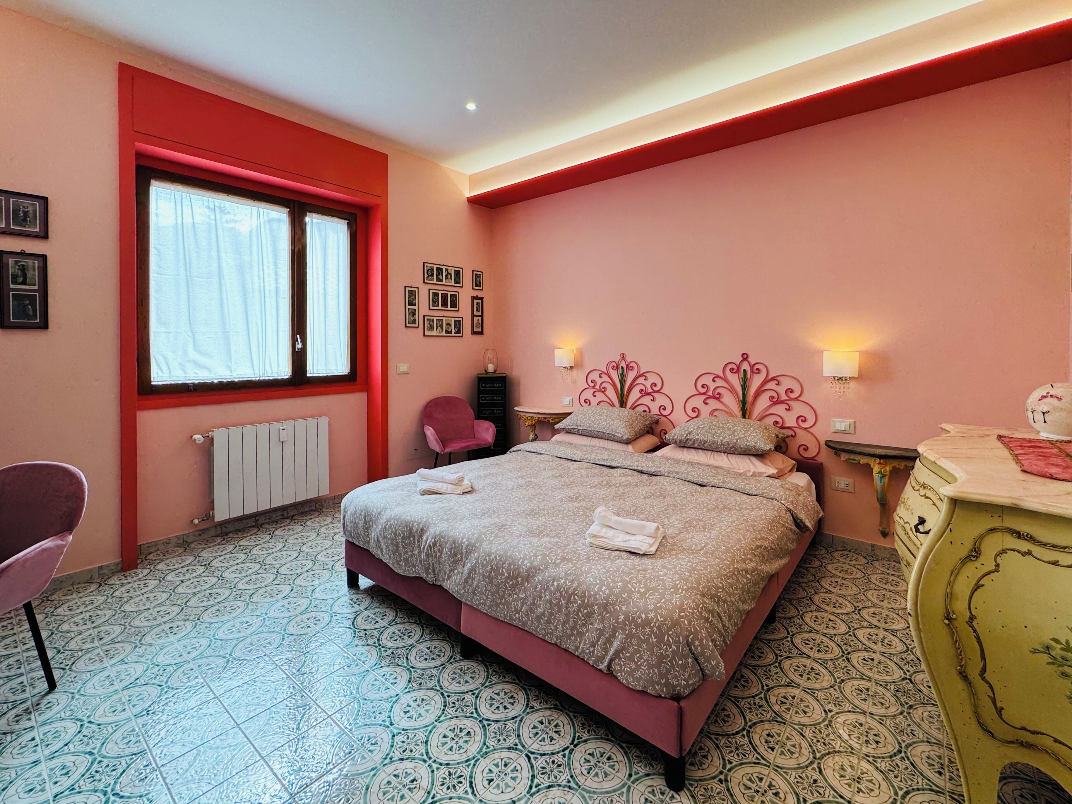 Un dormitorio con cama, una silla, cómoda, radiador, ventana e iluminación en la pared.