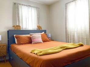 Una cama con cabecera, dos almohadas, una colcha y una mesita de noche con lámpara.