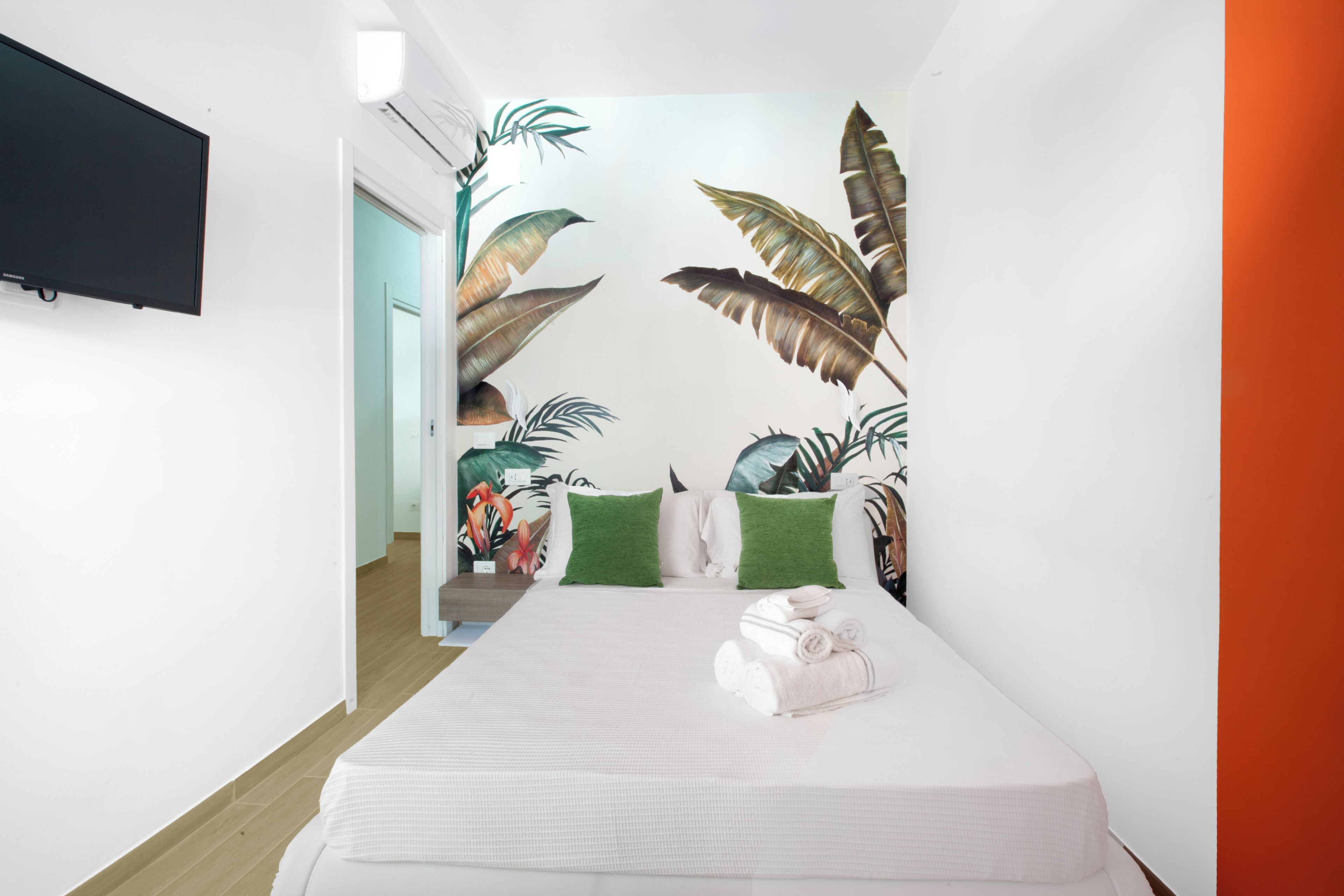 A hotel room with a bed, green pillows, a flat-screen TV, and a tropical wallpaper.